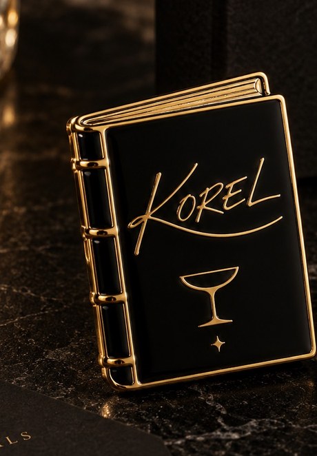 KOREL Volume I enamel pin cover, a small black and gold book charm engraved with the KOREL wordmark and a coupe glass